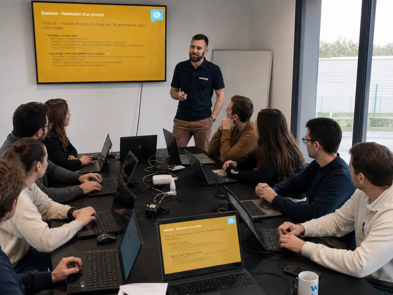 Formateur Wavia debout devant un groupe en formation IA, ordinateurs ouverts, l'exercice « Réalisation d'un prompt » affiché à l'écran